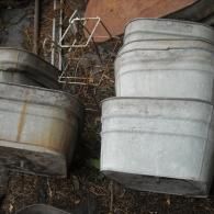 A stack of four rectangular galvanized metal wash tubs placed on the ground alongside a wire rack.
