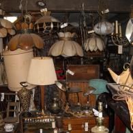 A cluttered antique shop filled with hanging pendant lamps, table lamps with fabric shades, and various vintage decor.