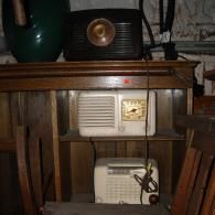 Three vintage radios sit on a wooden shelf, with a dark, rounded object visible above them.