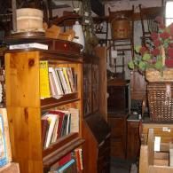 A wooden bookshelf filled with books, standing in a cluttered shop or antique store filled with furniture and baskets.