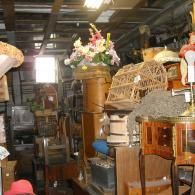 A crowded, cluttered antique shop interior filled with furniture, a wicker birdcage, and decorative floral arrangements.