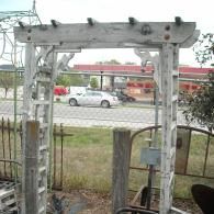 A weathered, light-colored wooden garden trellis stands outdoors with an open view of a street and distant buildings.