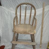 A rustic, weathered wooden child's high chair with a spindle back and footrest, positioned in front of a white drape.