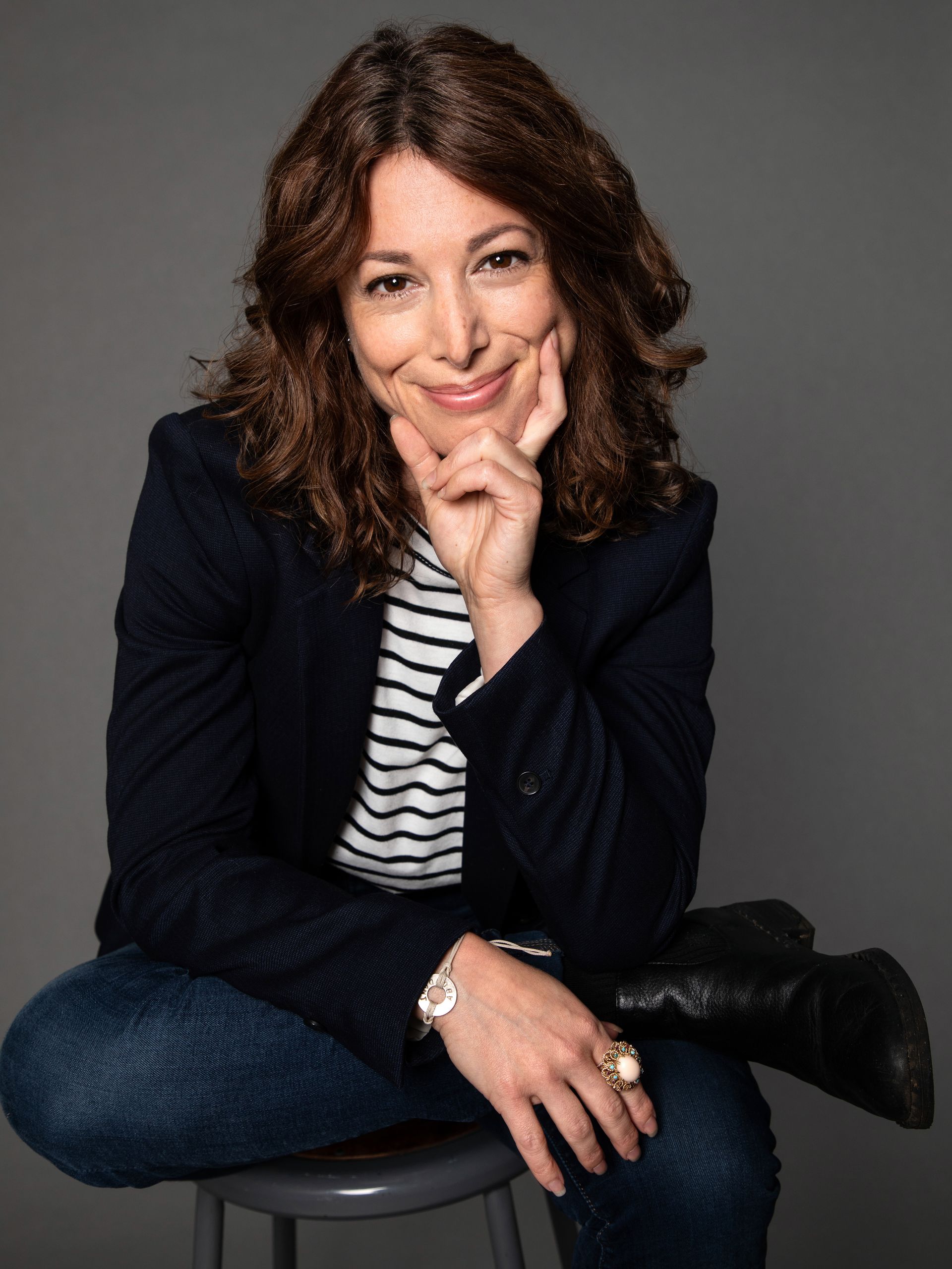 Image description: a woman with shoulder length curly brown hair sits on a stool against a gray background. Her right ankle rests on her left knee as she smiles warmly, chin in her hand and looks directly at the camera. She's dressed in a black blazer, with a black and white striped T-shirt, jeans and black ankle boots. She wears a large ring and bracelet. 