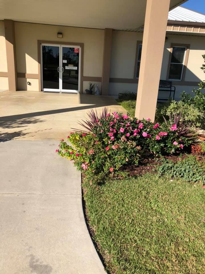 A building entrance with glass doors, a tan support column, and a garden of pink flowers and green grass.