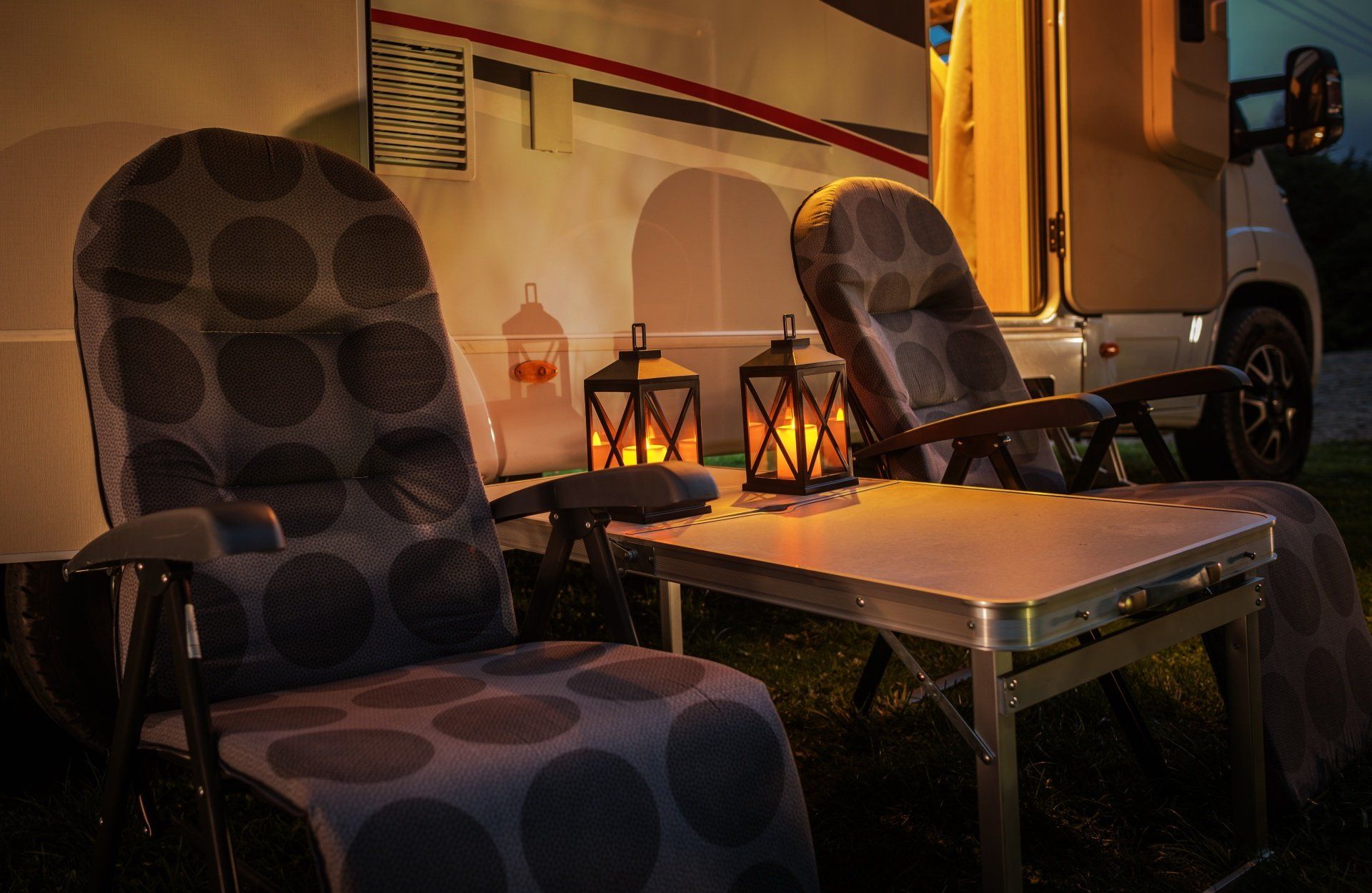 Two chairs with polka dot cushions, a small table, and lanterns lit in front of a motorhome at night.