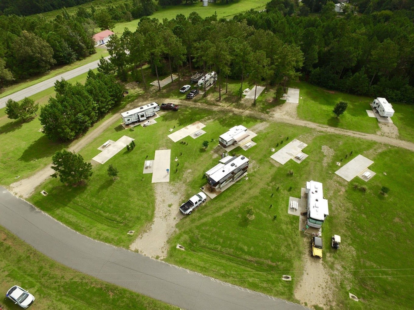 Aerial view of an RV park with several RVs parked on grassy lots, surrounded by trees and a paved road.