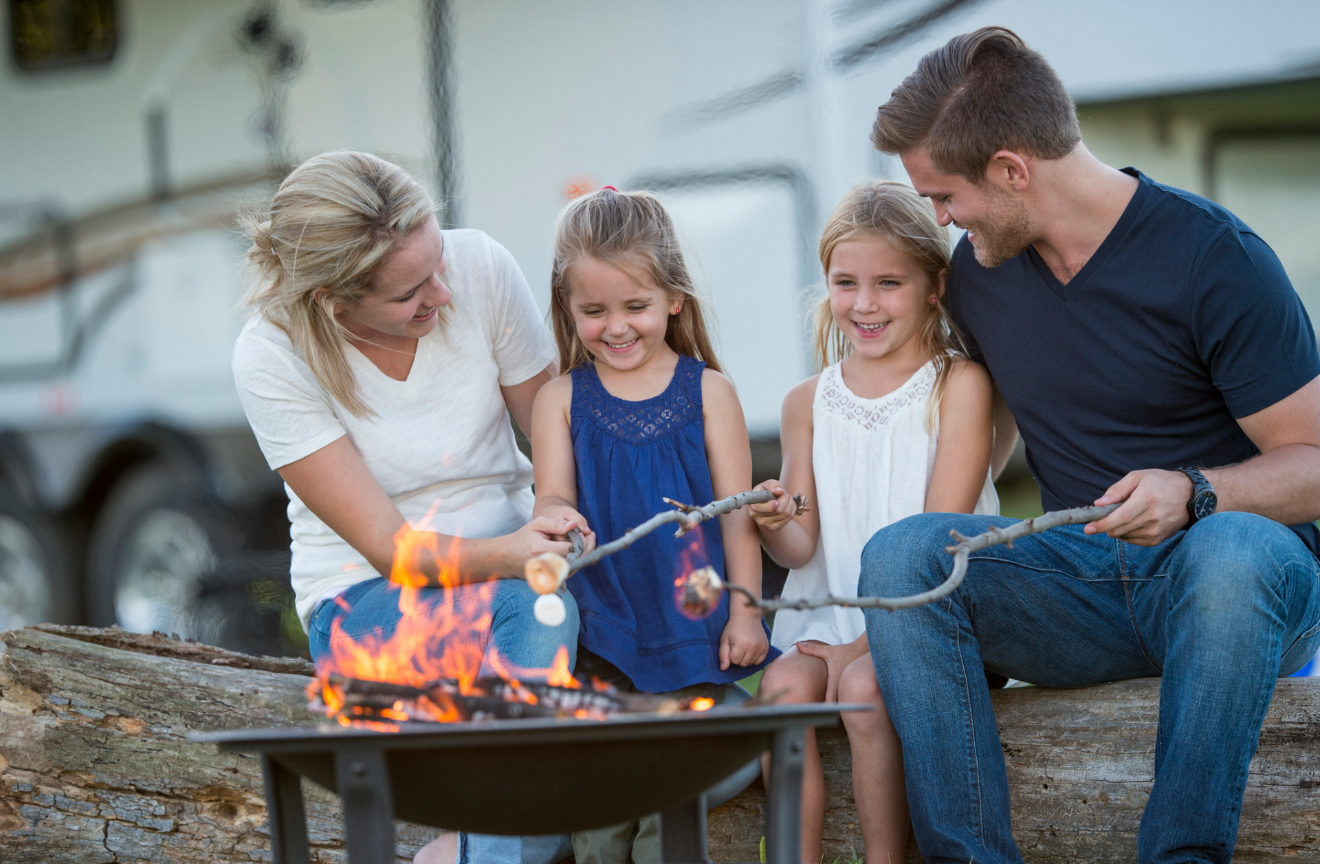 Family of four smiling and roasting marshmallows by a campfire near their white RV.