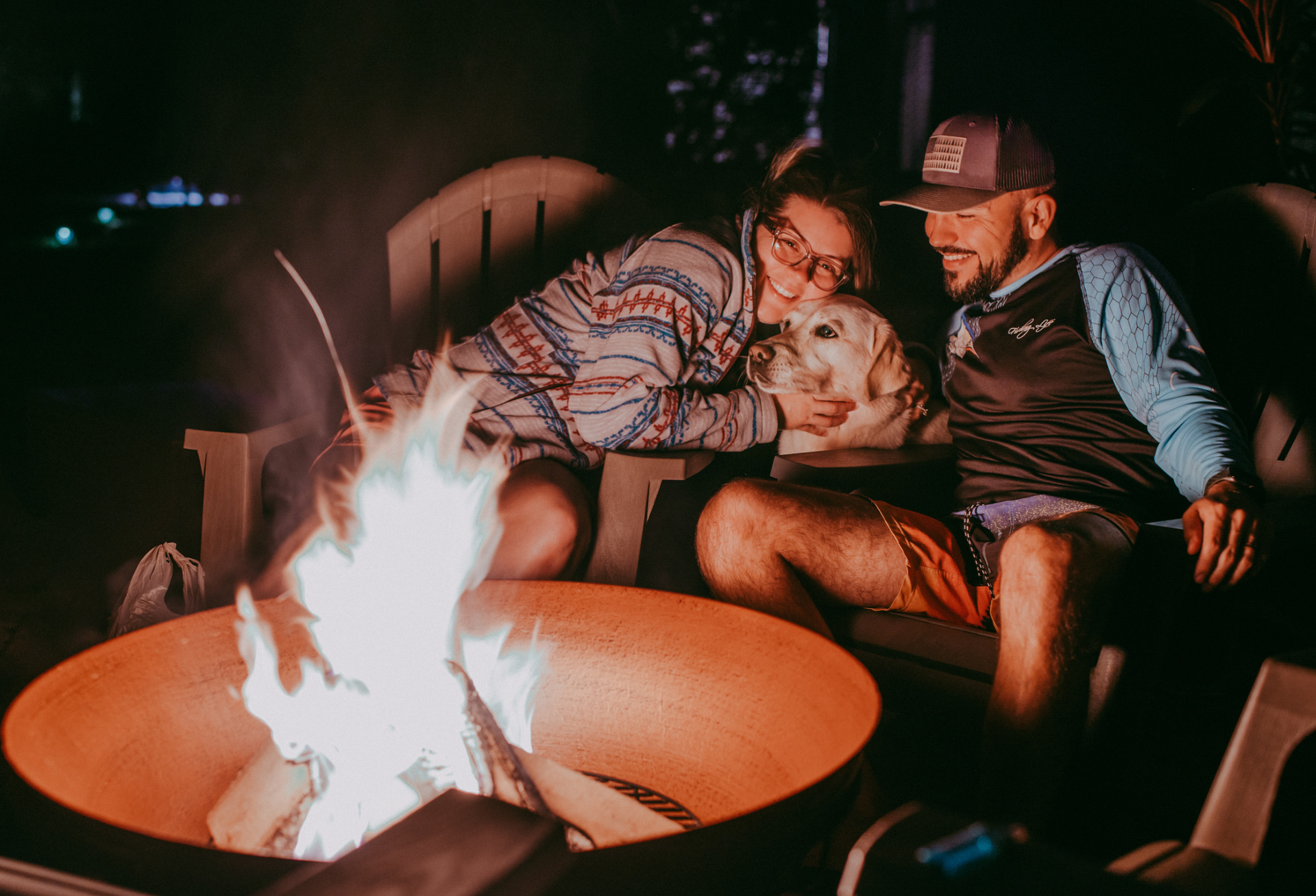 Couple and dog by fire pit at night, smiling.
