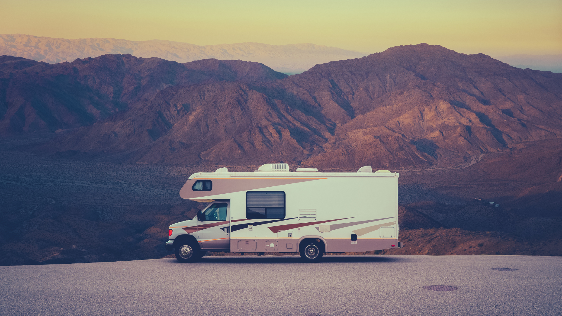 RV parked on a road with mountain backdrop at sunset. Beige and white vehicle, brown mountains.