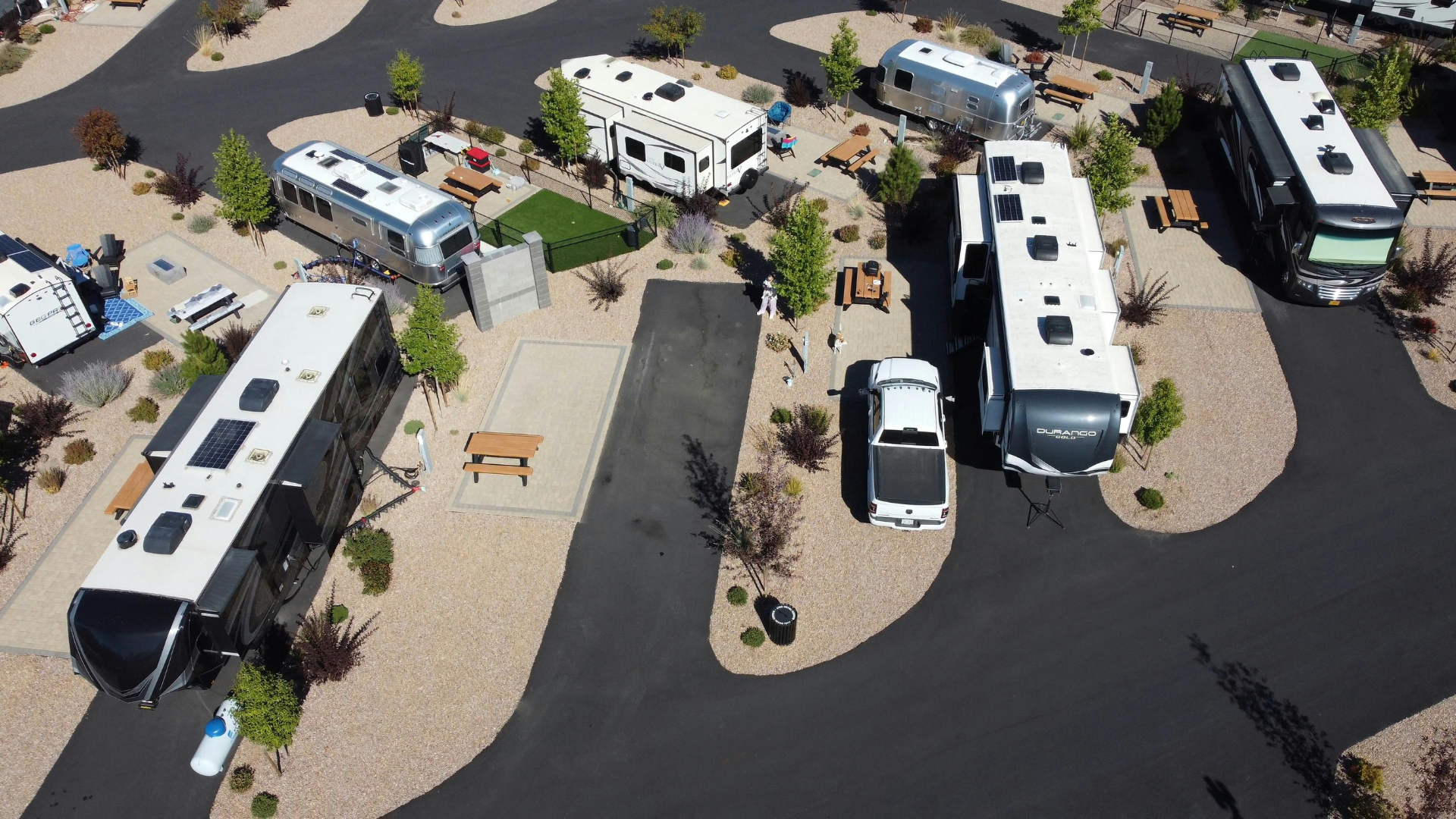 Aerial view of an RV park with various RVs parked on asphalt pads surrounded by gravel, trees, and roads.