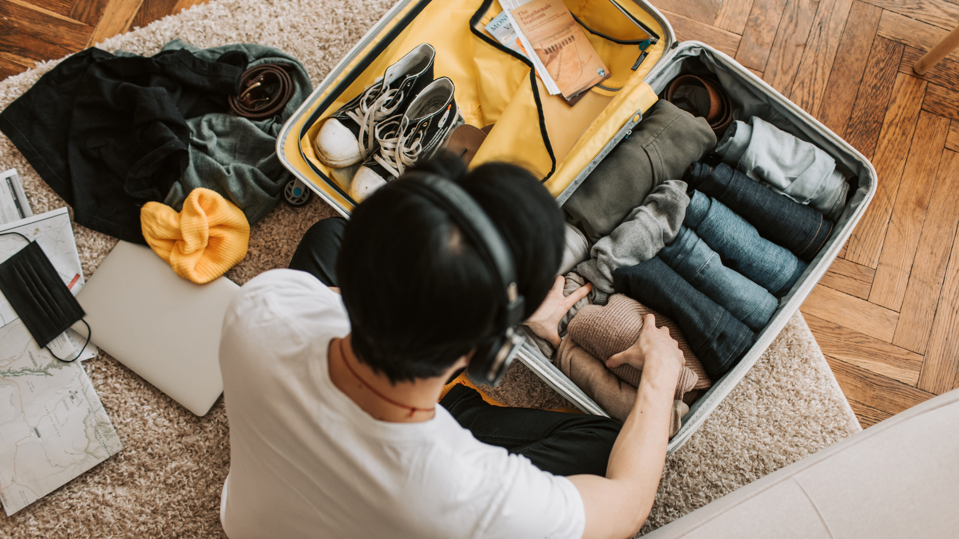 Person packing a suitcase with clothes, headphones, and laptop on the floor.