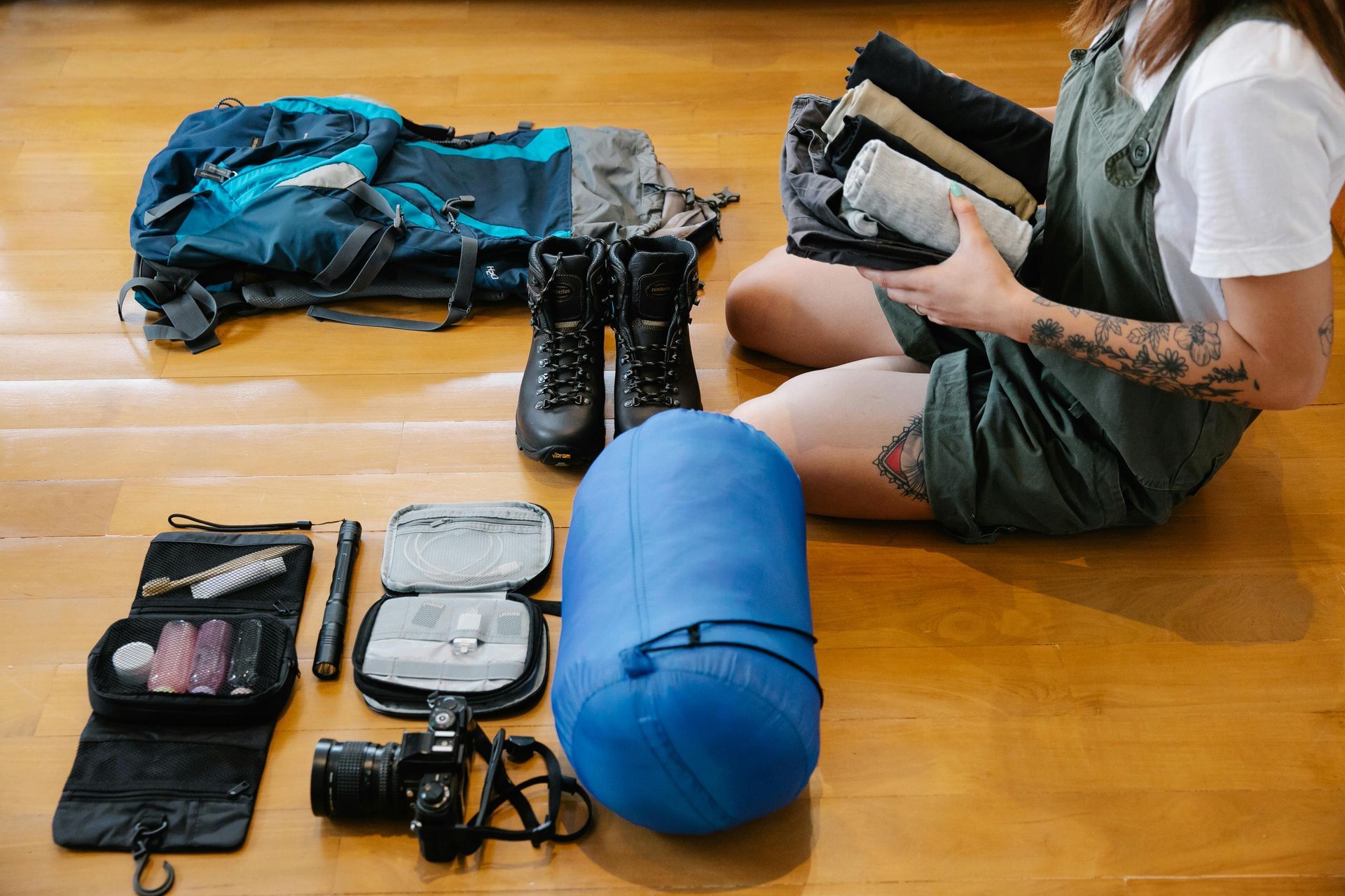 Hiking boots, a rolled blue sleeping bag, a camera, and a survival kit on a table.