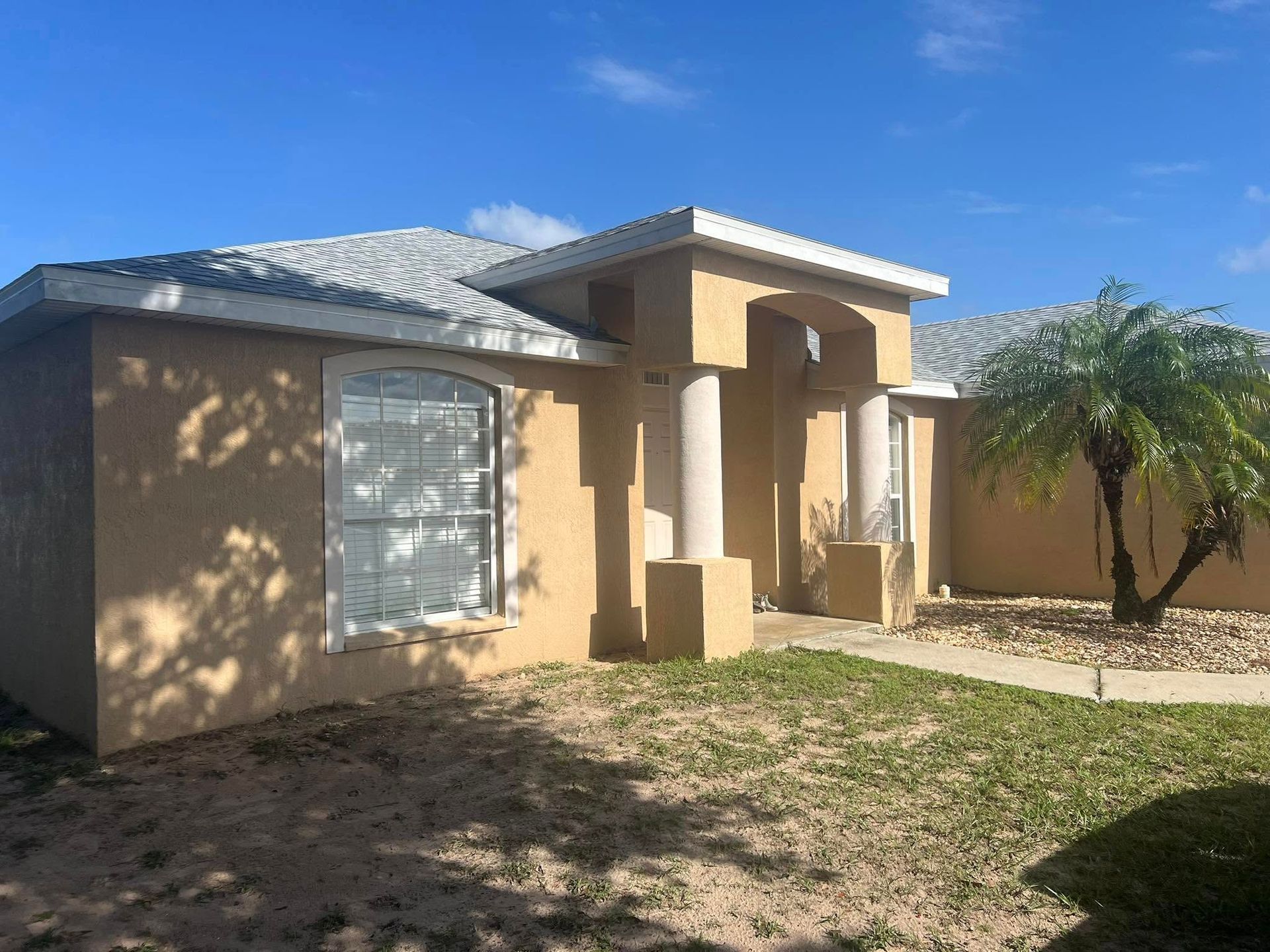 Tan stucco house with white trim under a bright blue sky. Entrance has a portico supported by pillars.