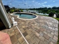 Poolside patio with a pool, statue, and view of a lake under a blue sky. The patio is made of brown pavers.