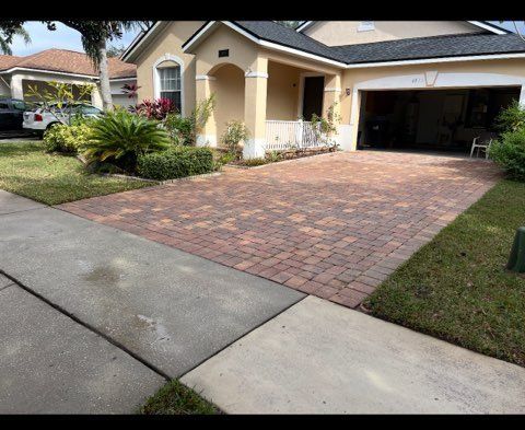 Brick paver driveway in front of a beige house with a two-car garage; green lawn and sidewalk.