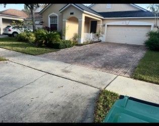 A house with a brick paved driveway and a green recycling bin in the foreground, suburban setting.