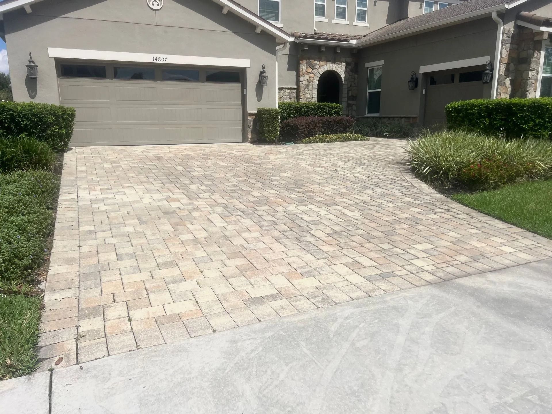Brick driveway leading up to a two-car garage of a stucco house, with manicured hedges and green grass.