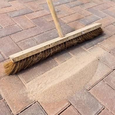 A broom sweeps sand into the gaps between brick pavers, outdoors.