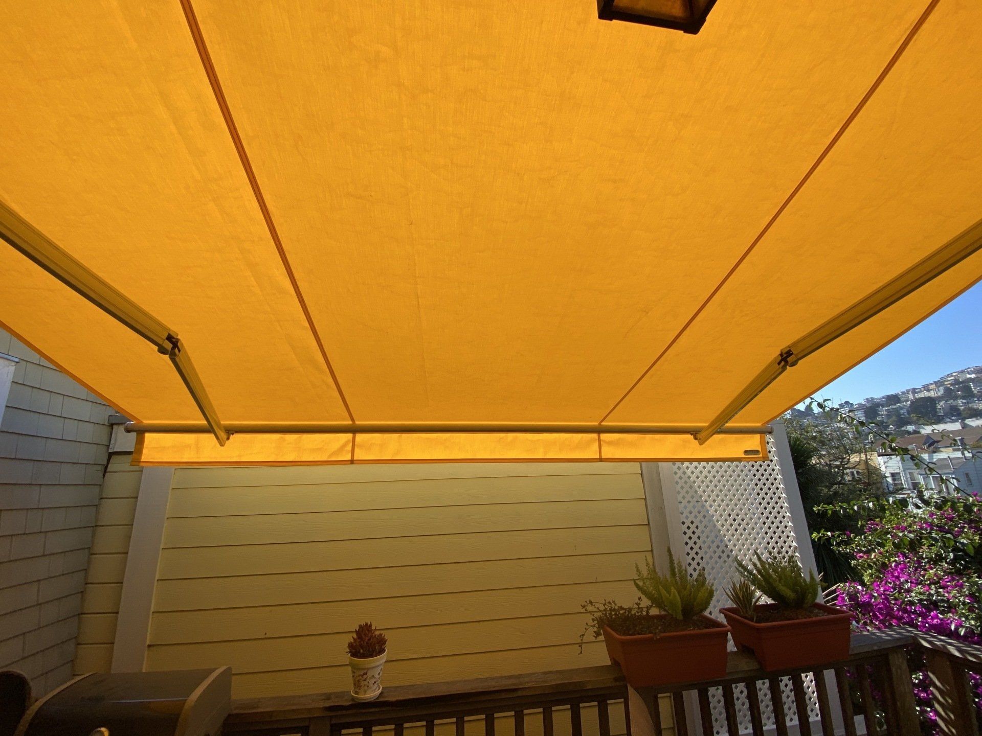 A yellow awning is covering a balcony with potted plants