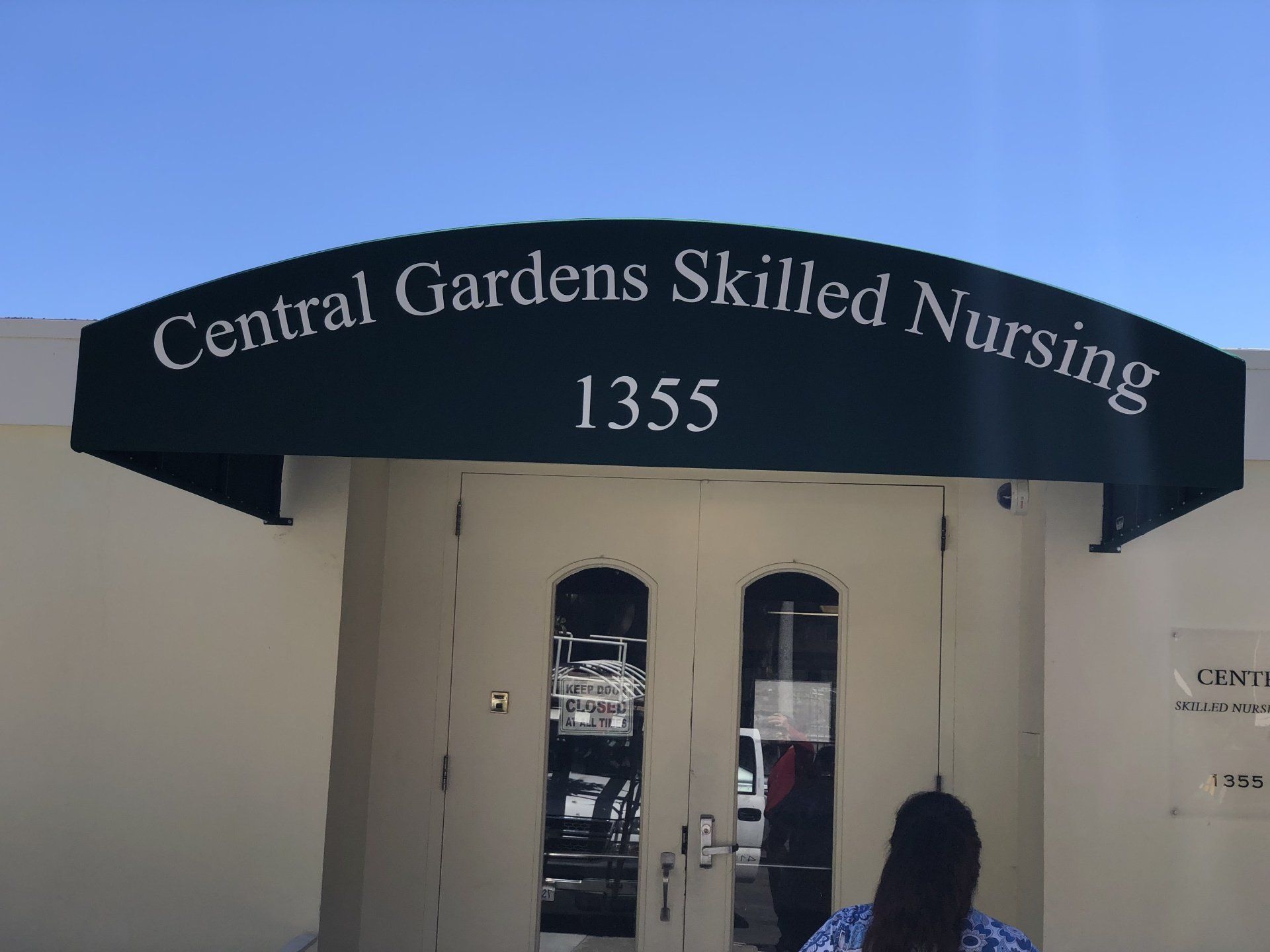 A woman stands in front of the central gardens skilled nursing building