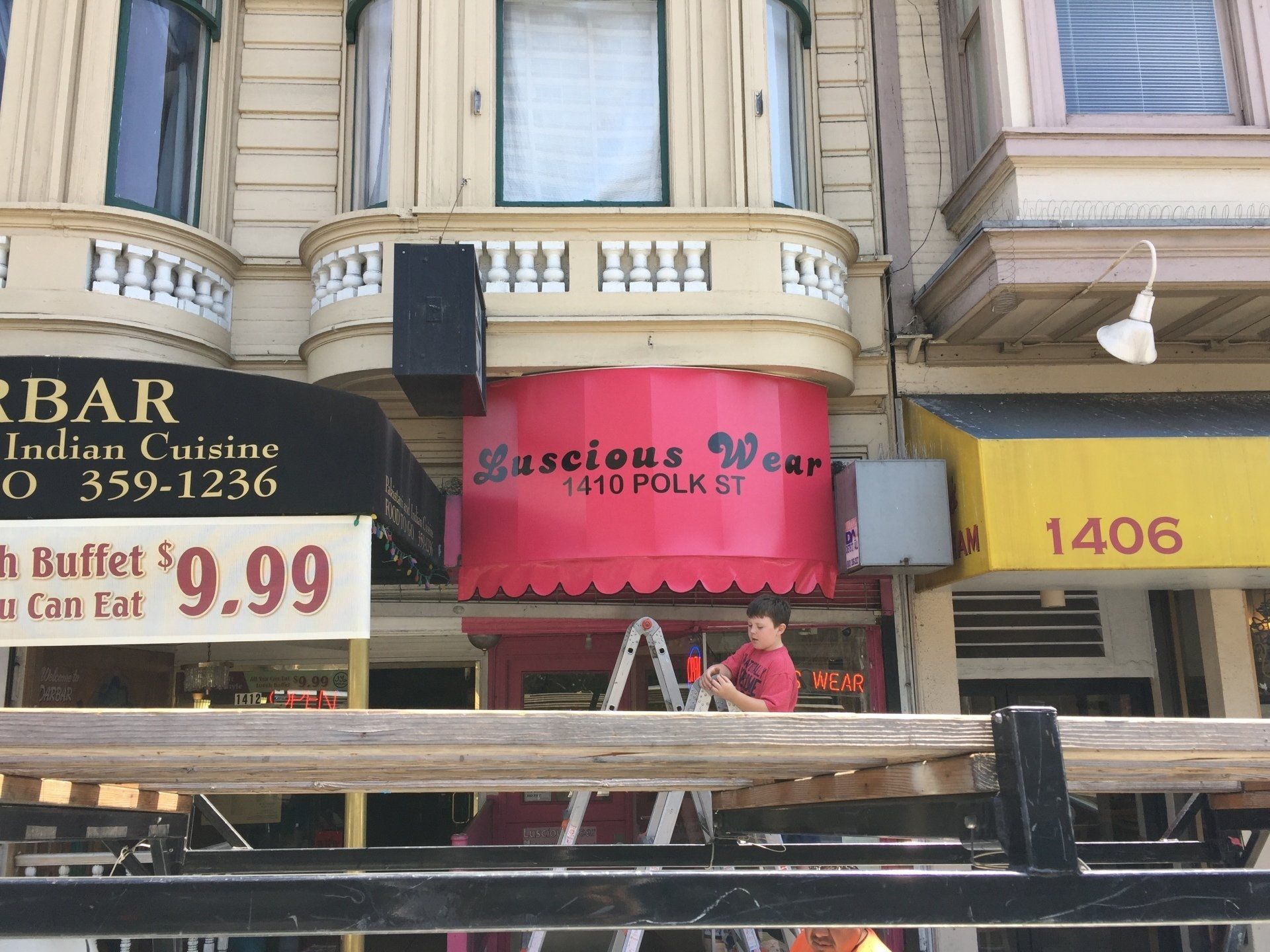 A man stands on a ladder in front of a red awning that says ' luscious wear ' on it