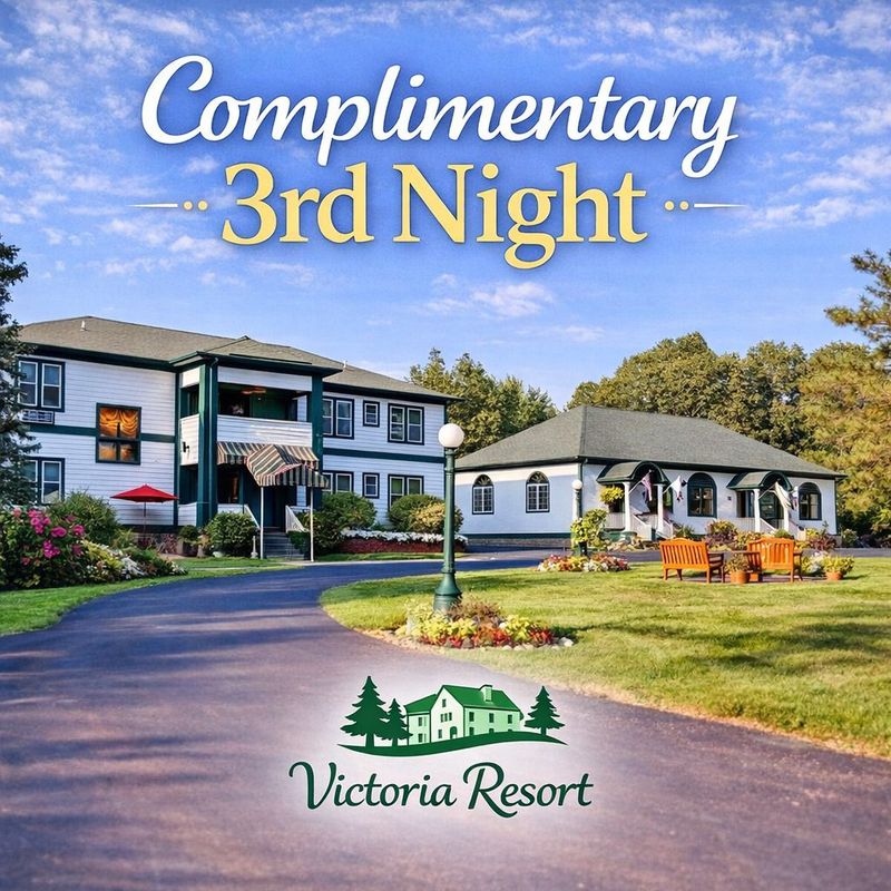 Two-story white building with green trim, benches on a brick patio, green grass, and trees under a blue sky.
