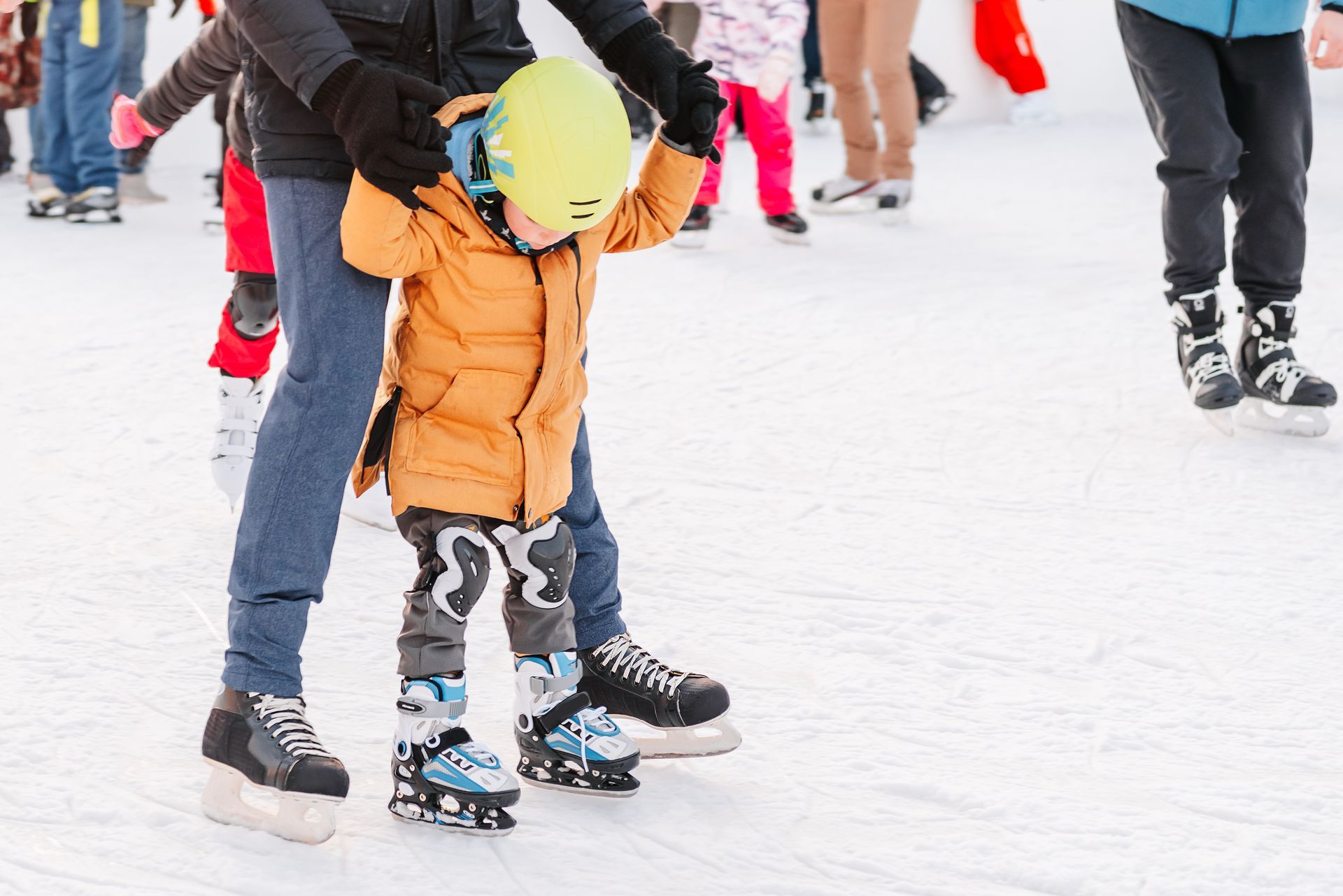 Person helping a child ice skate outdoors; the child wears a yellow helmet and orange jacket.