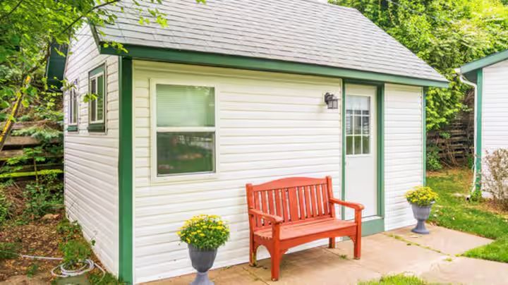 White shed with green trim, door, window, red bench, and potted plants on a concrete patio.