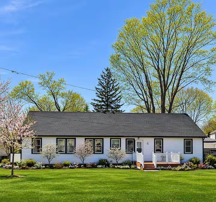 White cottage with green roof and deck, lush green foliage and pink flowers in foreground.
