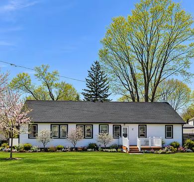White cottage with green roof and deck, lush green foliage and pink flowers in foreground.