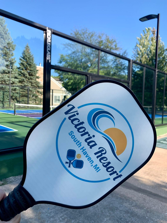 Basketball court with hoop, green and red playing surface, chain link fence, and trees.
