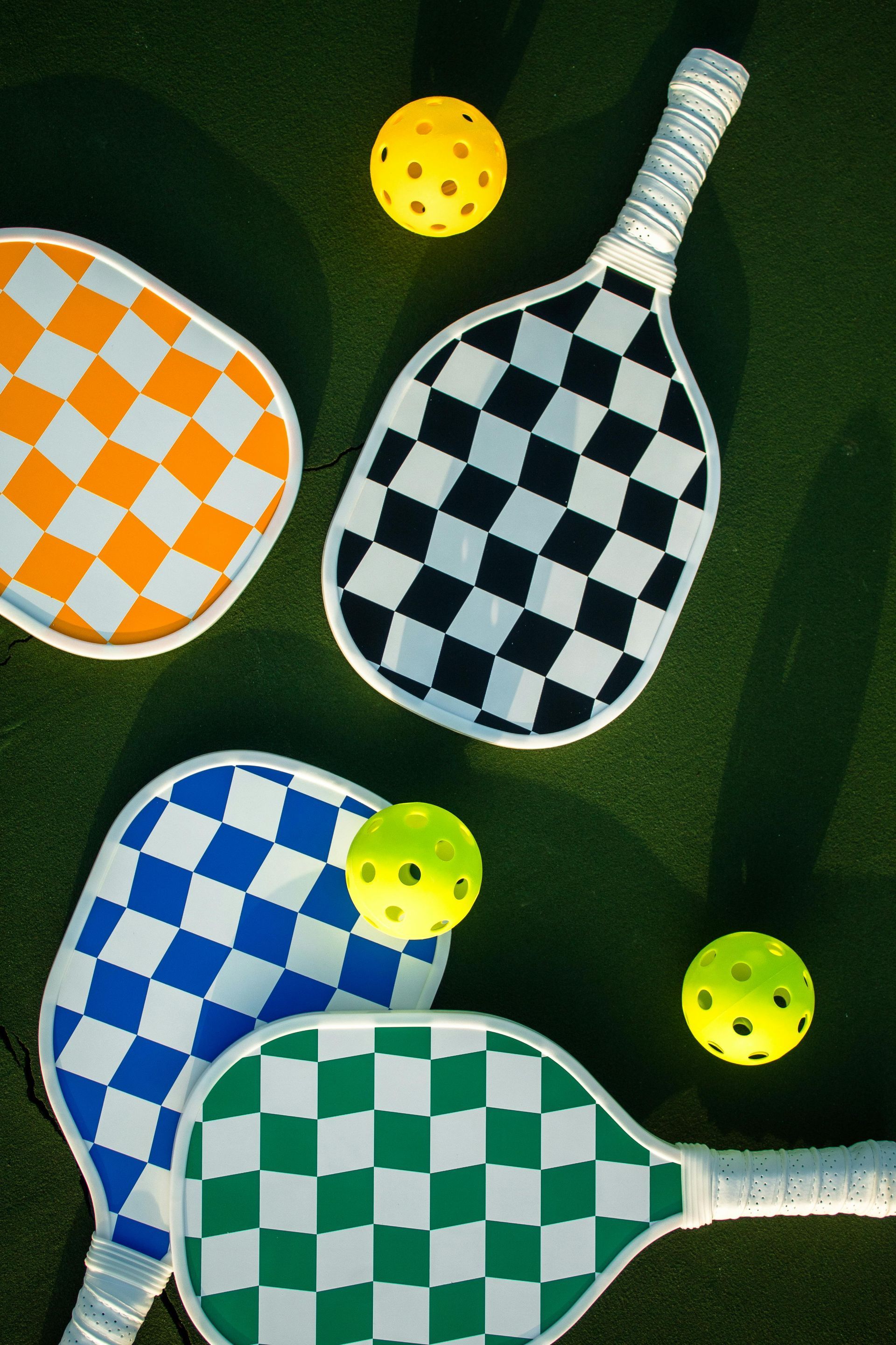 Pickleball paddles (orange, black, blue, green) and yellow balls on a green court.
