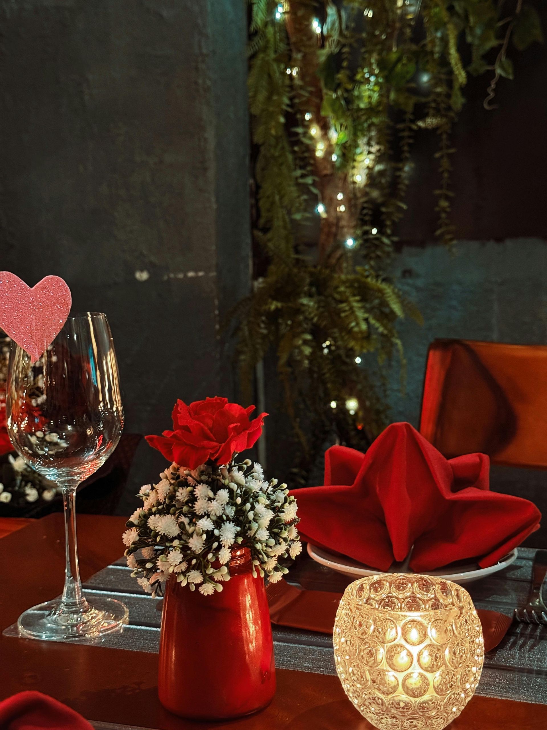 Romantic dinner setting: red rose centerpiece, wine glass, folded napkin, and candle on a table.