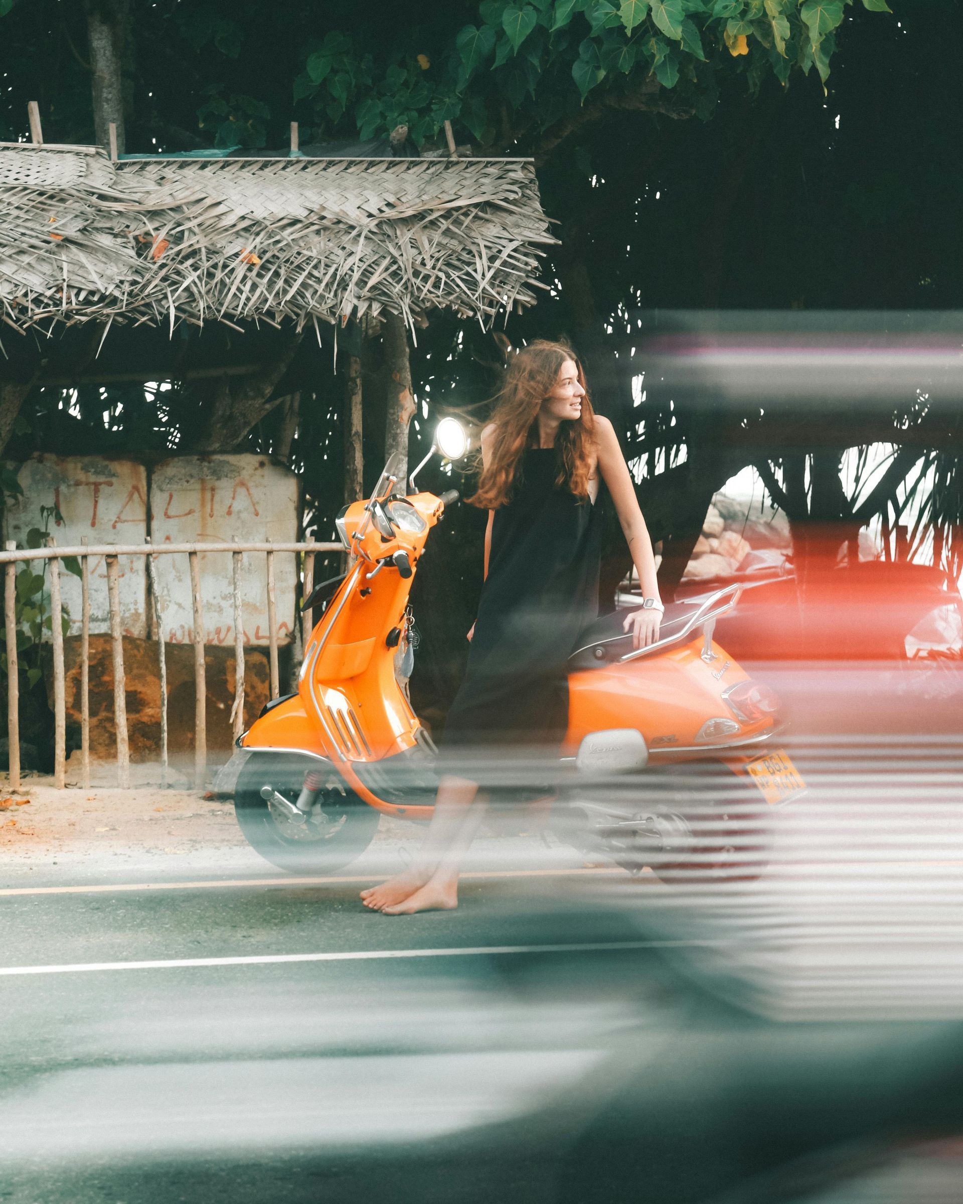 Woman on orange scooter at a crosswalk, blurry motion. Bare feet, black dress.