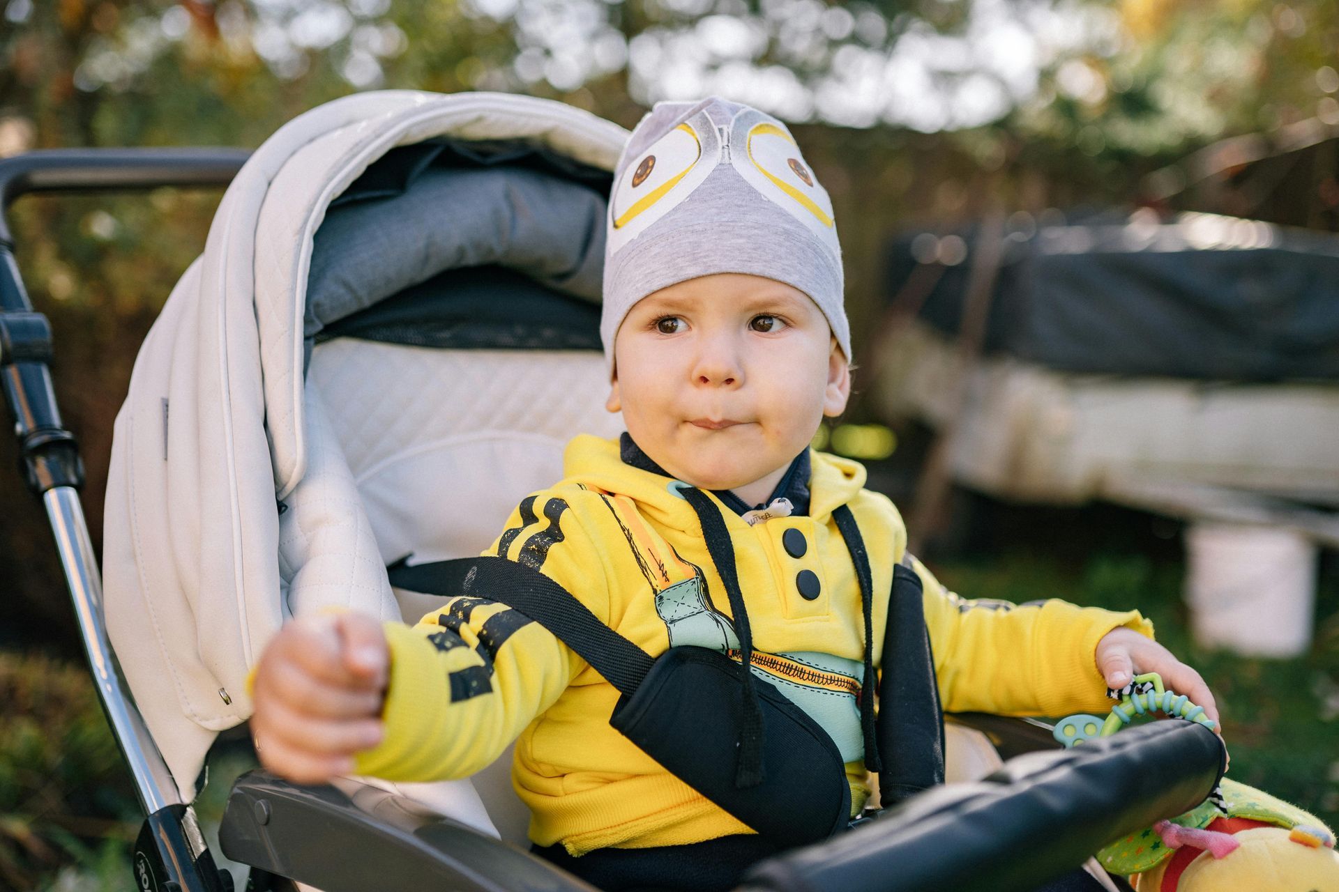 Child in a stroller wearing a yellow jacket and owl hat outdoors.