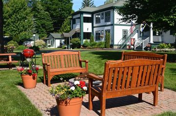 Wooden benches and potted flowers on a brick patio in front of a white building with green trim.