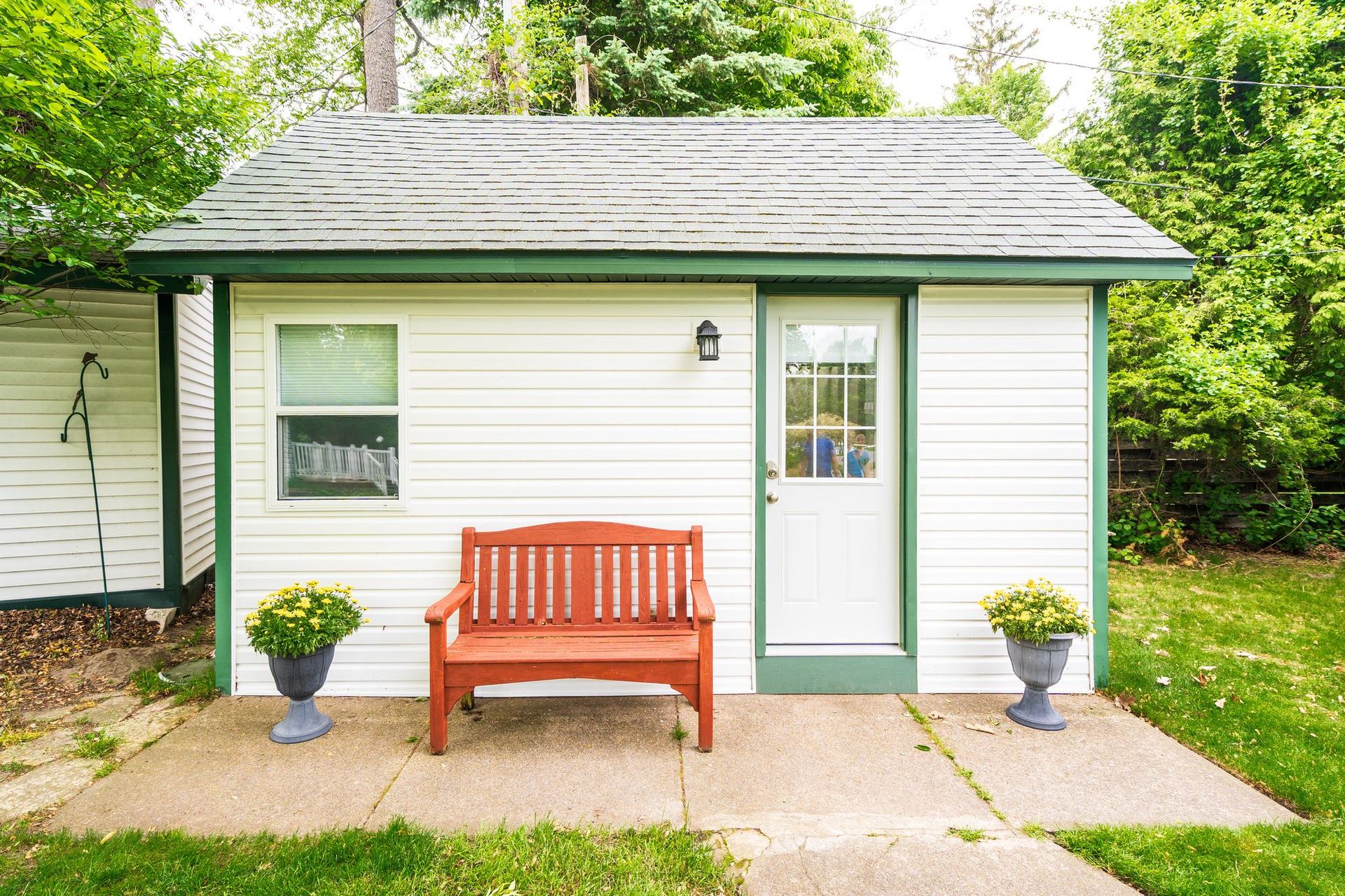 White shed with green trim, a red bench, two potted plants, and a small window.