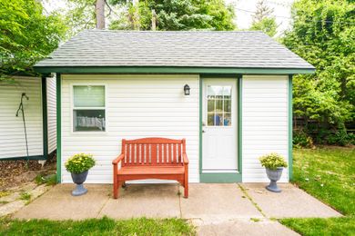 White shed with green trim, door, window, red bench, and potted plants on a concrete patio.