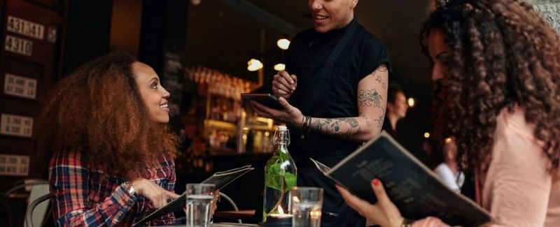 A server taking orders from two women at a restaurant. One woman reads a menu.