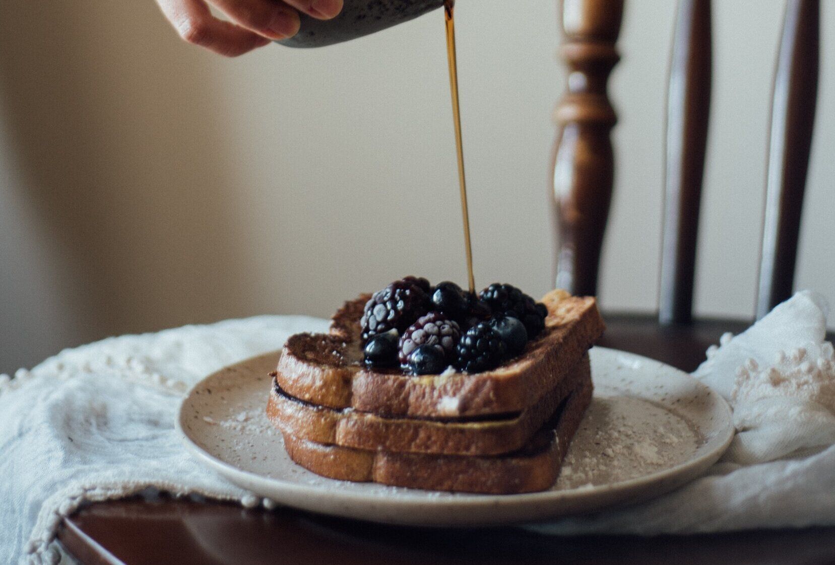 Maple syrup being poured on a stack of french toast topped with blackberries on a plate.