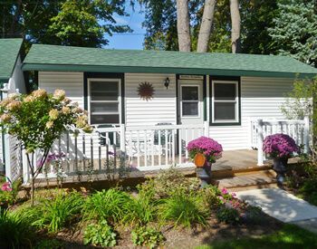 White cottage with green roof and small porch, surrounded by a garden of flowers.