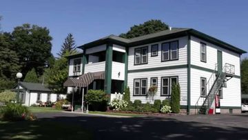 Two-story white building with green trim, awning, and fire escape. Sunny day, trees in background.