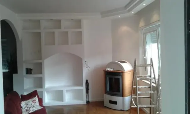 Scaffali bianchi incorporati e camino in una stanza con pavimento in legno e finestra; è presente una scala.