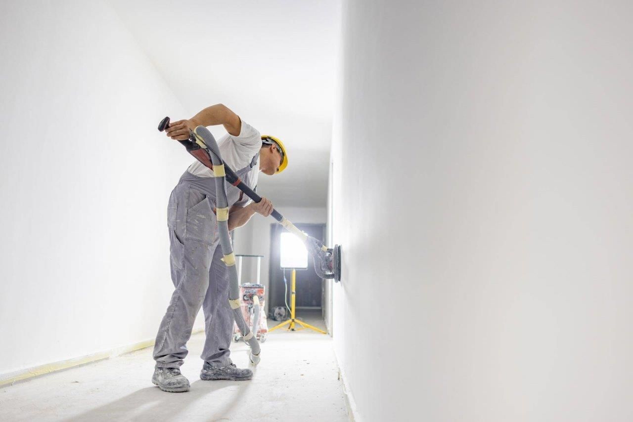 Un operaio edile carteggia una parete bianca con un lungo utensile abrasivo in un corridoio vuoto.