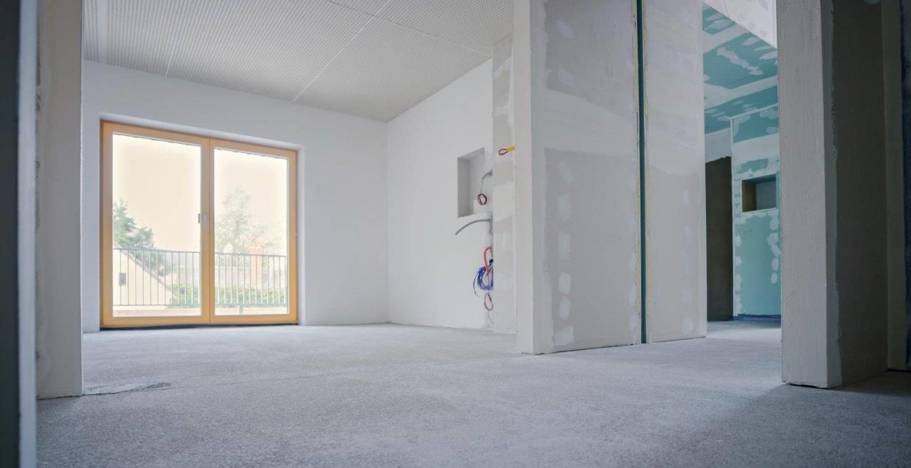 Stanza vuota in costruzione con pareti bianche, cartongesso grezzo, ampia finestra e moquette grigio chiaro.