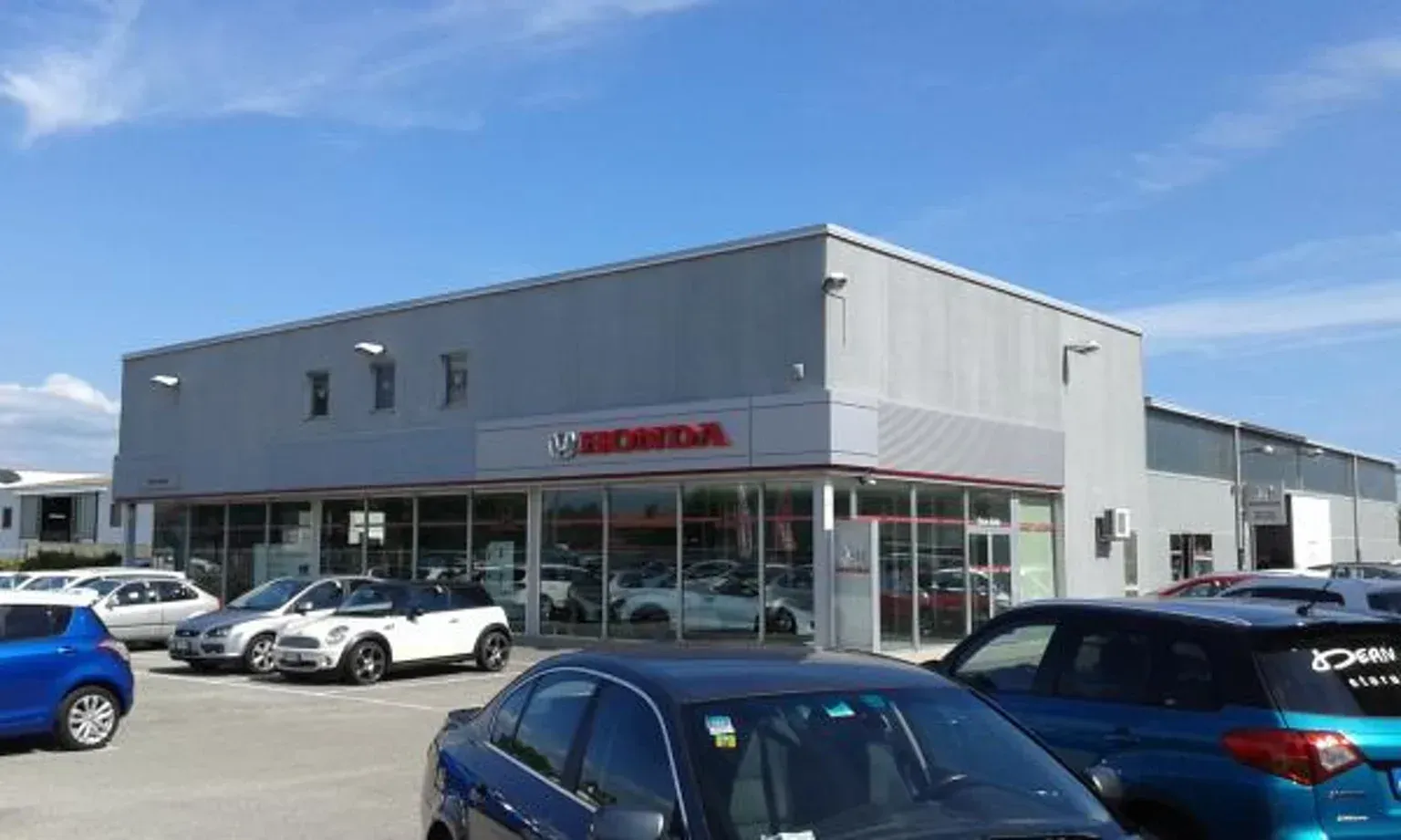 Concessionaria Honda con auto parcheggiate di fronte. Edificio grigio, logo Honda rosso, cielo azzurro.