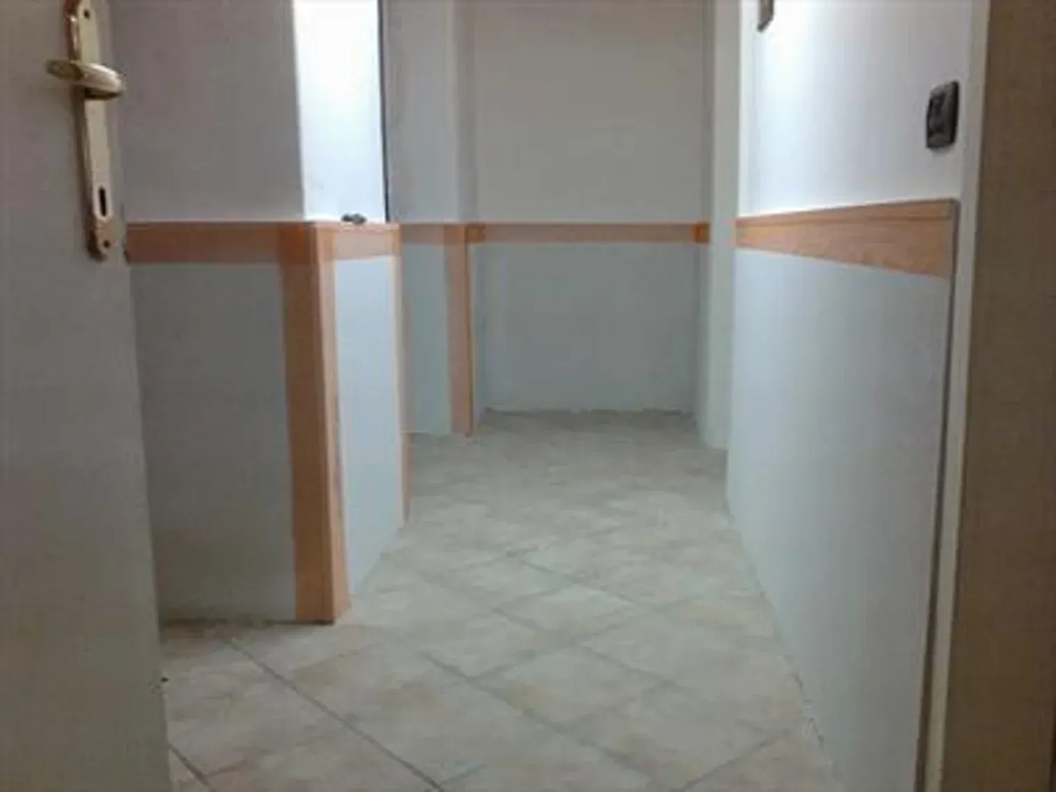 Corridoio vuoto con pareti bianche, finiture color cuoio e pavimento piastrellato. Porta sulla sinistra.