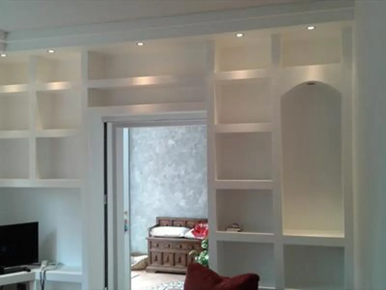 Mensole bianche integrate con illuminazione da incasso incorniciano una porta aperta, mobili a vista e una TV scura sulla sinistra.