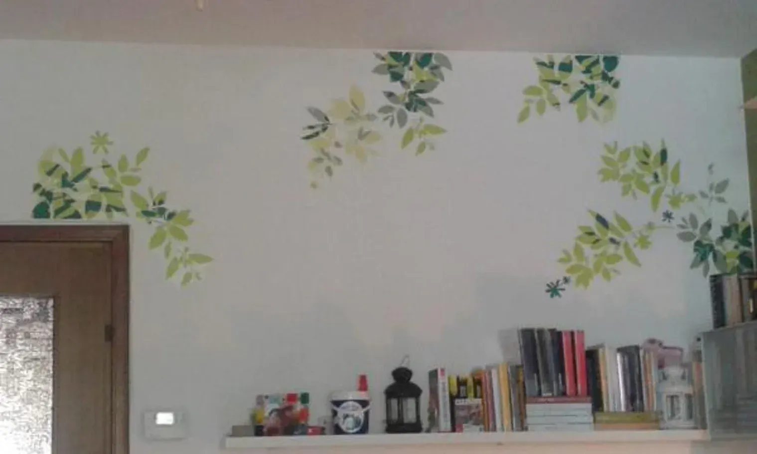 Parete bianca con motivi di foglie verdi dipinti, sopra uno scaffale con libri e una lanterna scura.